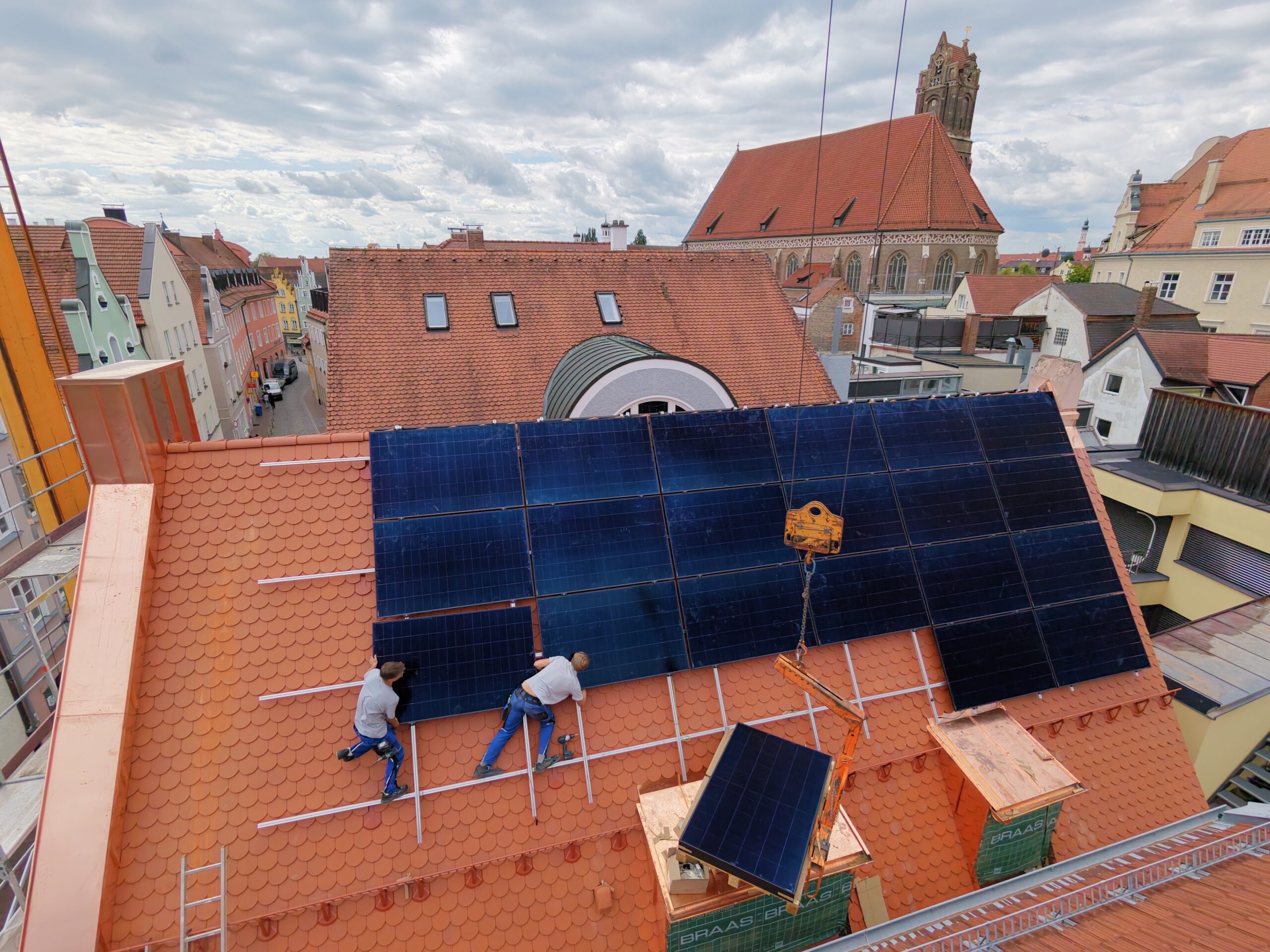 Zwei Monteure befestigen Solarmodule auf rotem Dach, Landshuter Martinskirche im Hintergrund