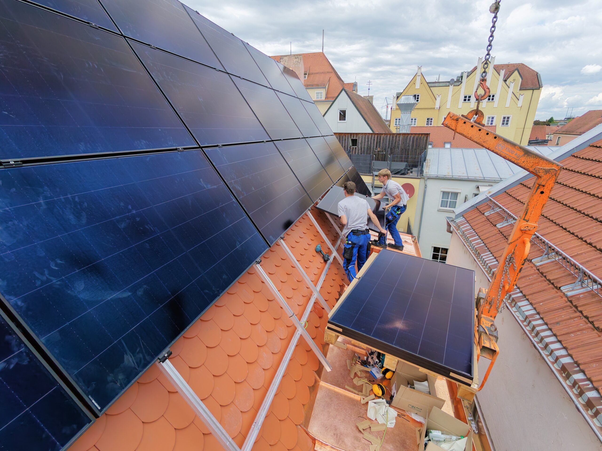 Zwei Handwerker installieren PV-Module auf Ziegeldach, Kranhaken über Altstadtdächern