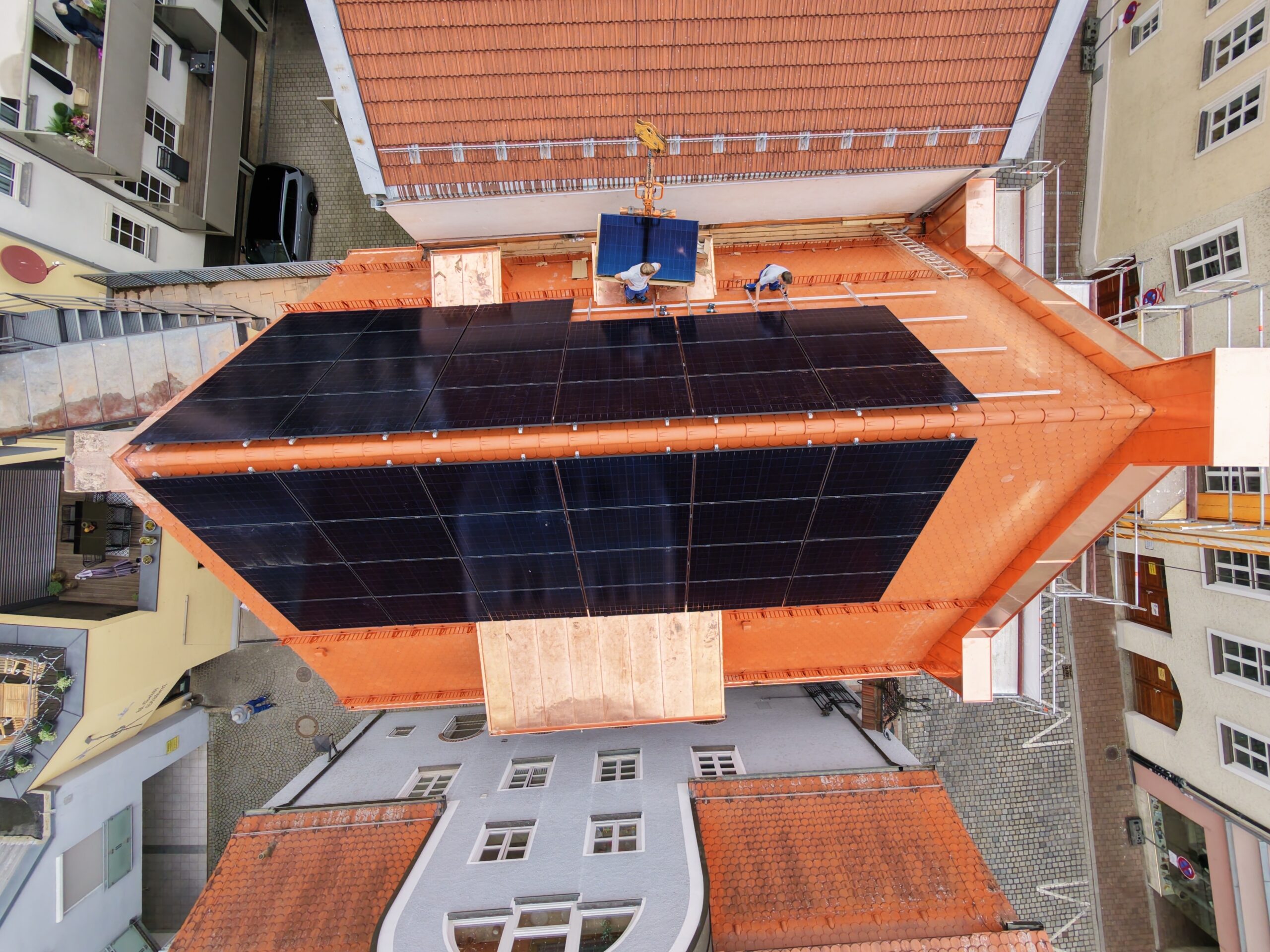 Luftaufnahme einer Photovoltaikanlage auf Altstadtdach, Monteure setzen per Kran ein Modul